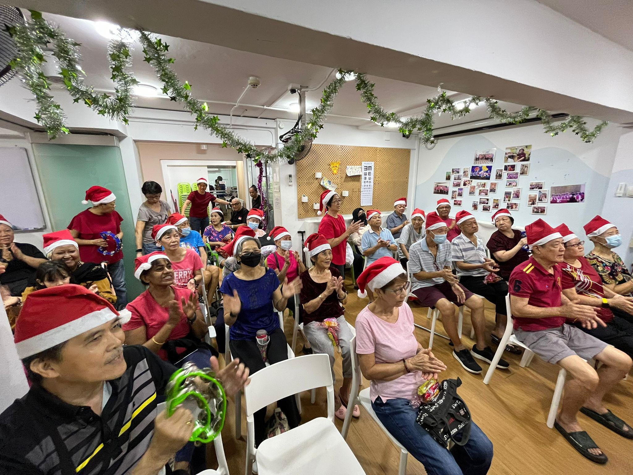 A group of adults wearing Santa hats sit in chairs, clapping and playing musical instruments, with Christmas decorations in the room.